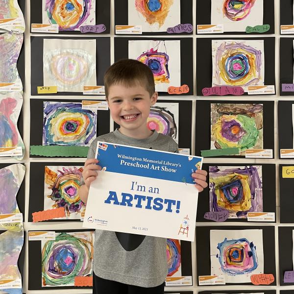 Child standing in front of artwork holding a sign that says, 